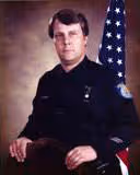 Portrait of a man in a black police uniform seated with an American flag in the background.
