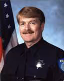 Police officer in uniform with badge and American flag in the background.