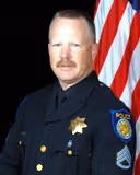 Police officer in uniform with a mustache standing in front of an American flag.