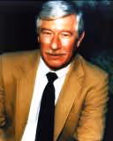 Portrait of a man with gray hair wearing a brown jacket, white shirt, and black tie against a dark background.