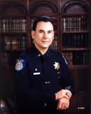 Portrait of a police officer in uniform sitting with hands clasped in a room with wooden bookshelves.