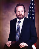 Man with beard and mustache wearing a dark suit and striped tie sitting in front of an American flag.