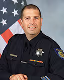 Uniformed male police officer smiling in front of a U.S. flag backdrop.