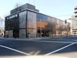 Modern glass and brick building on a street corner with reflections of surrounding trees and buildings.