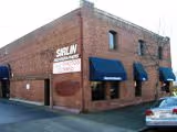 Brick building with blue awnings and sign reading 'SIRLUN' on corner near a parked white car.