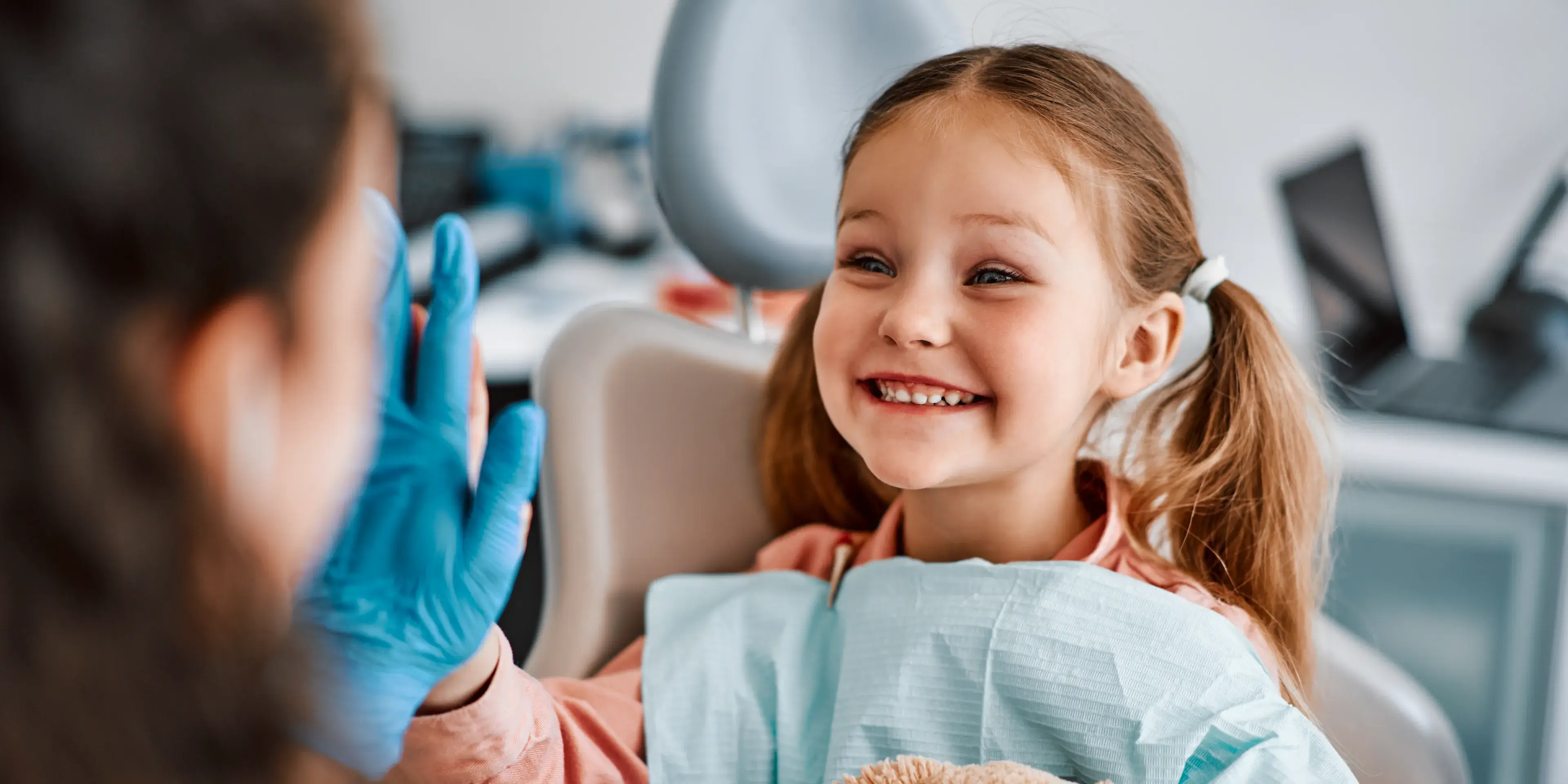 Smiling young girl in a dental chair giving a high five to a person wearing blue gloves.