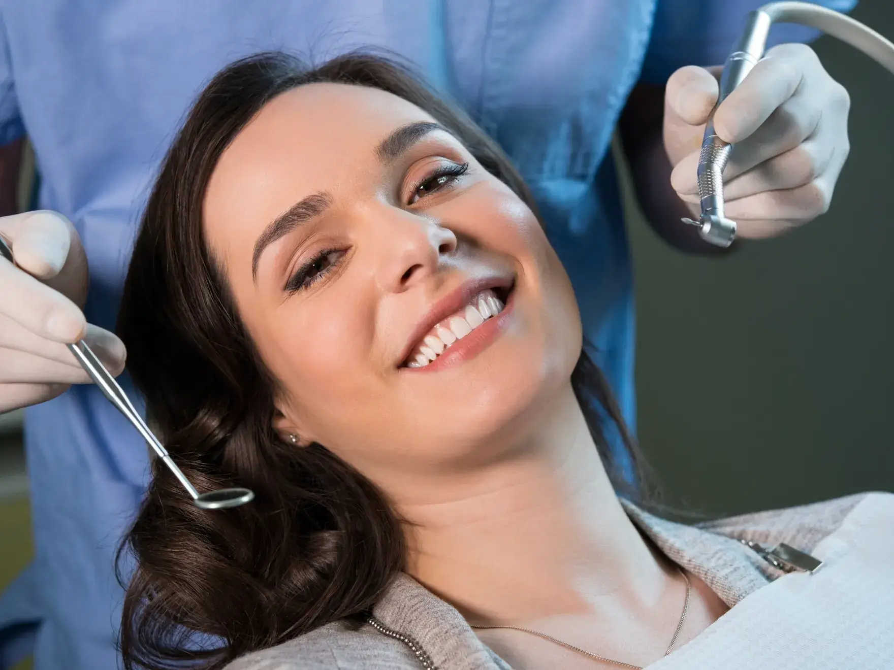 Smiling woman reclining in a dental chair with a dentist holding dental tools near her mouth.
