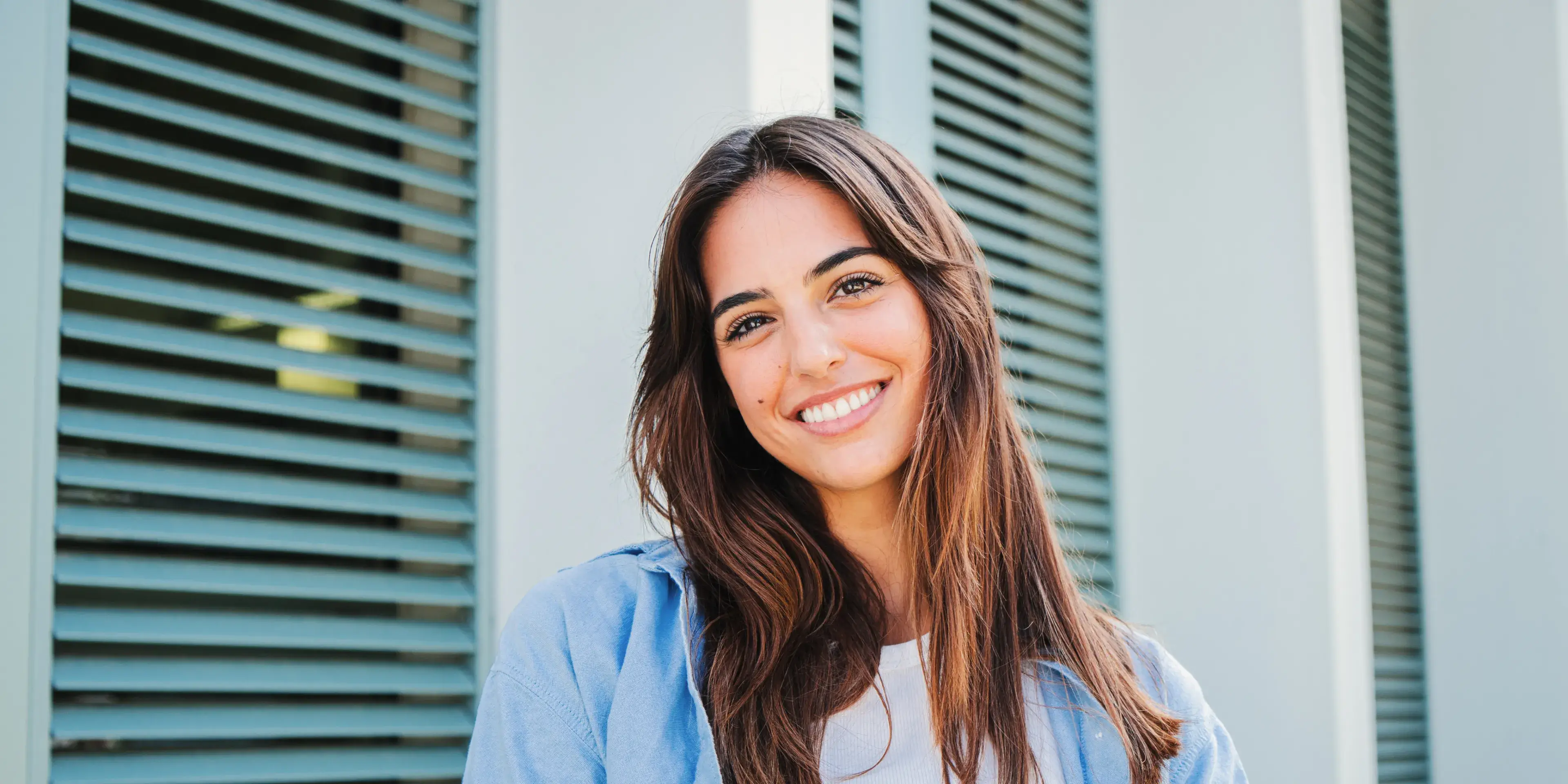 Smiling young woman with long brown hair wearing a light blue jacket in front of a building with blue shutters.
