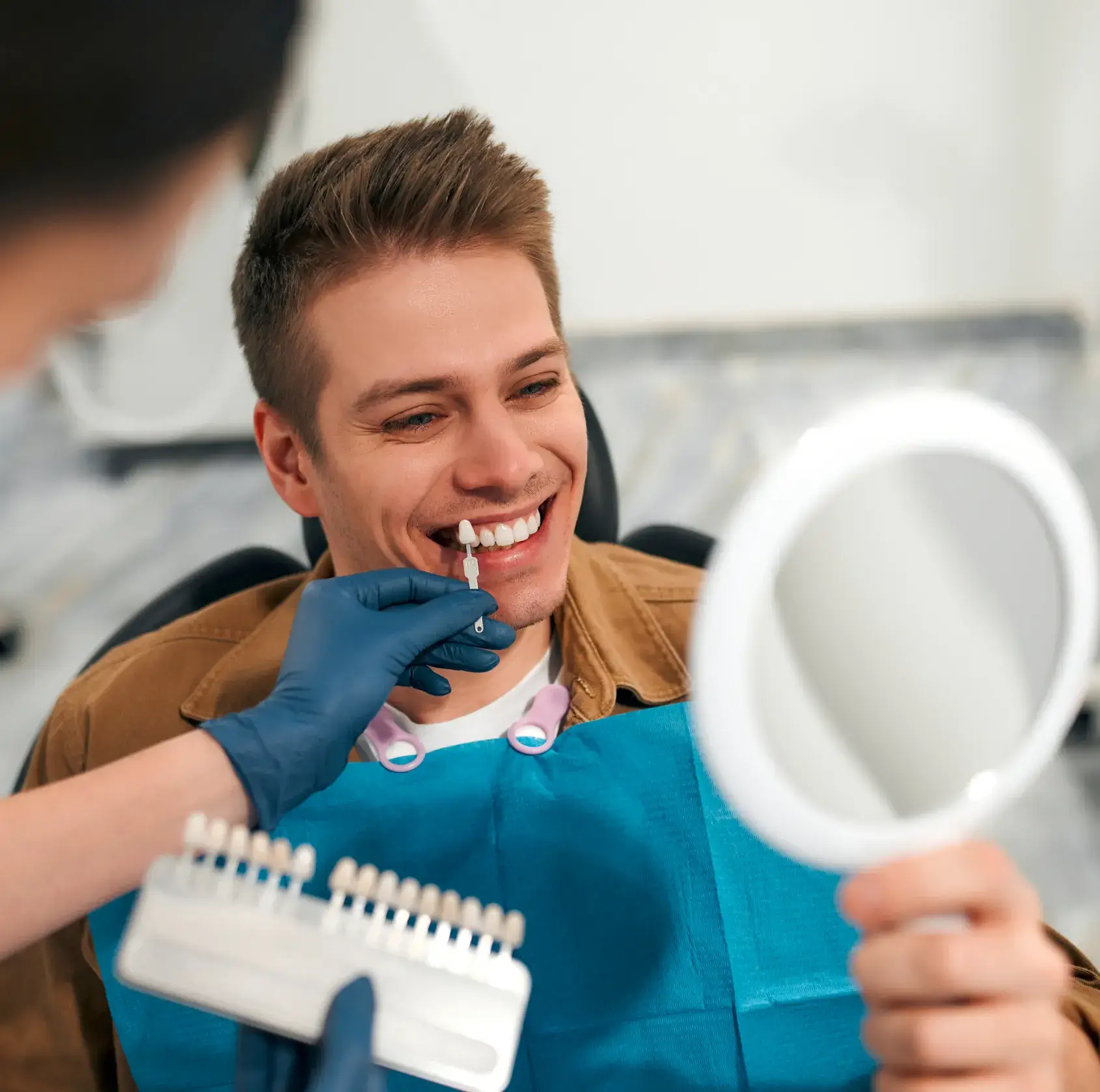 Dentist holding a dental shade guide near a smiling man's teeth while he looks in a handheld mirror.