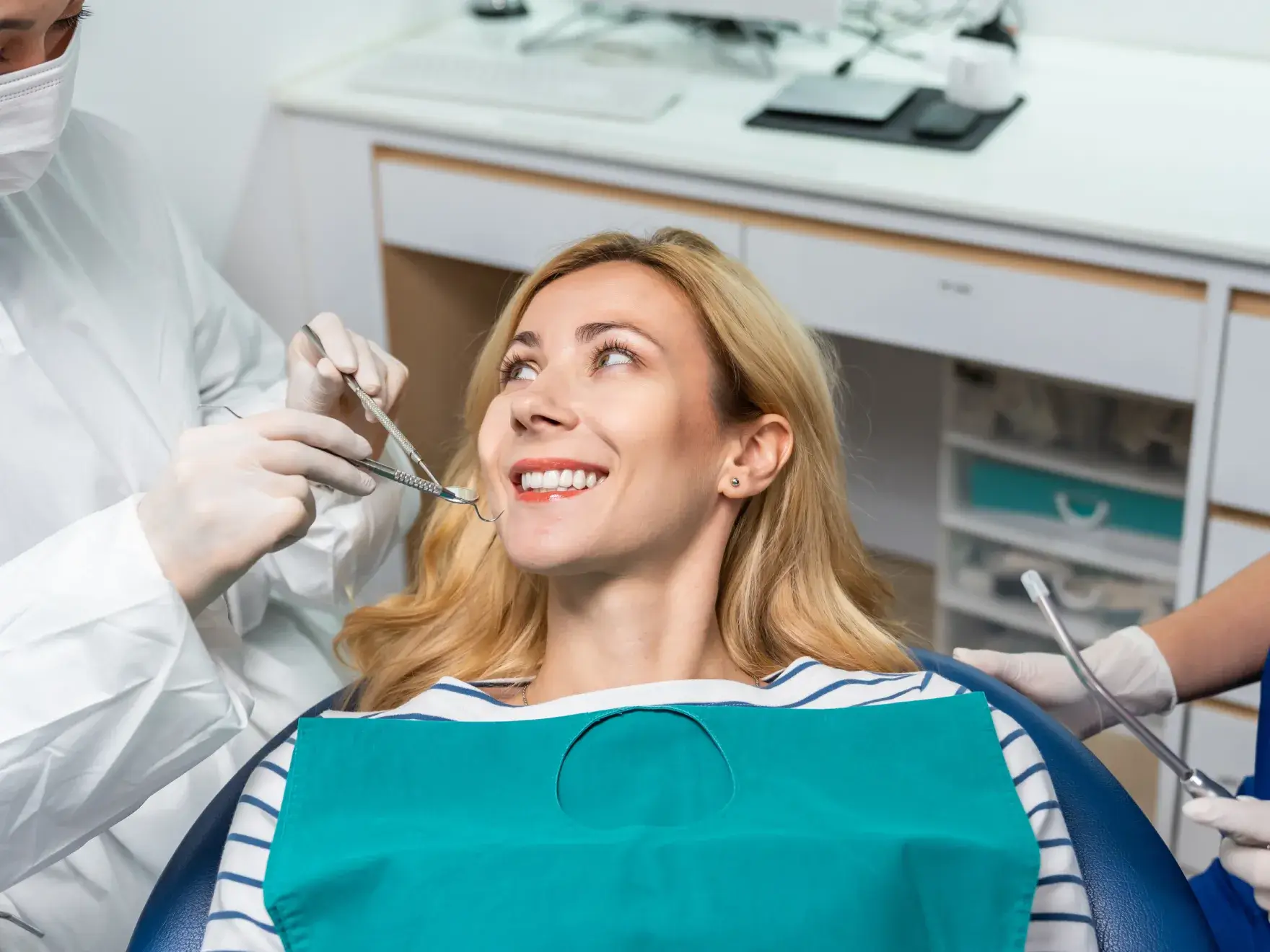 Smiling blonde woman in dental chair receiving dental checkup from masked dentist wearing gloves.