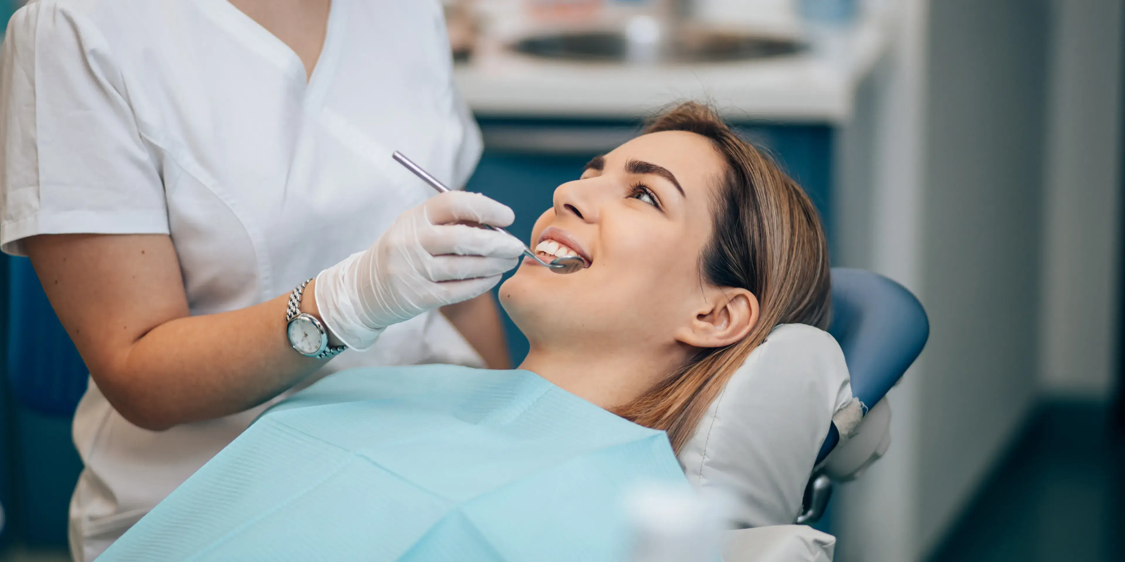 Smiling woman reclined in a dental chair while a dentist wearing gloves examines her teeth with dental instruments.