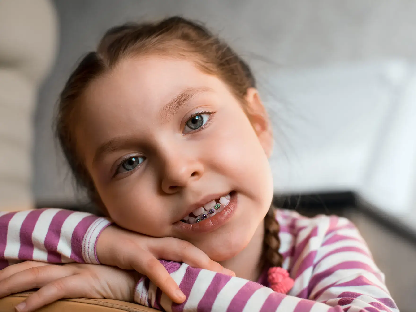 Smiling young girl with braces wearing a pink and white striped shirt resting her chin on her crossed arms.