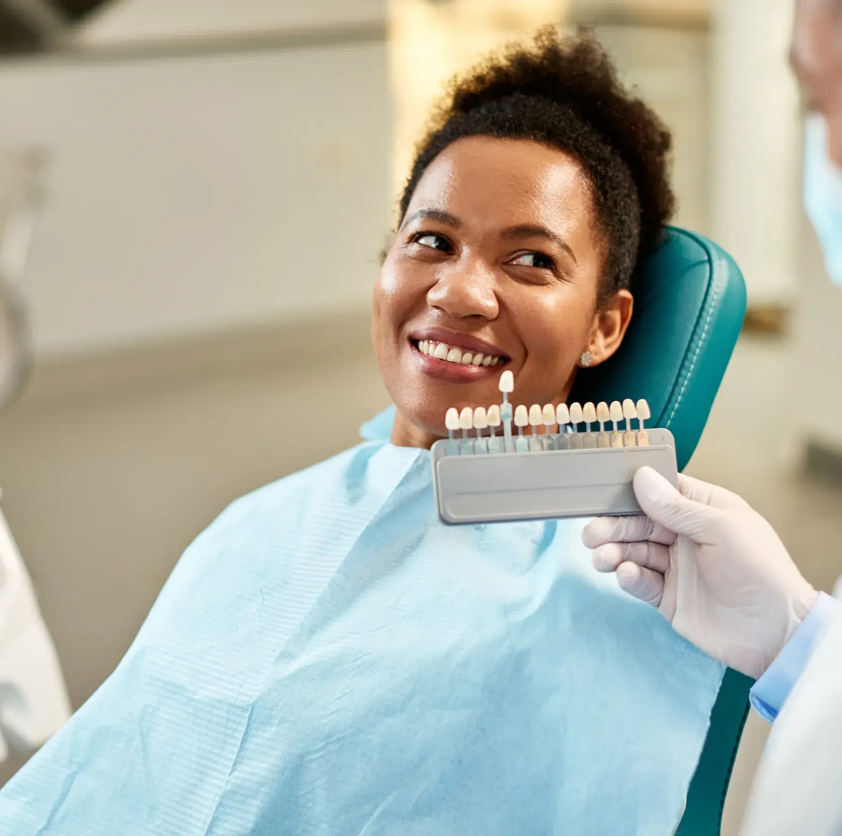 Smiling woman in dental chair comparing her teeth to a shade guide held by a gloved dentist.