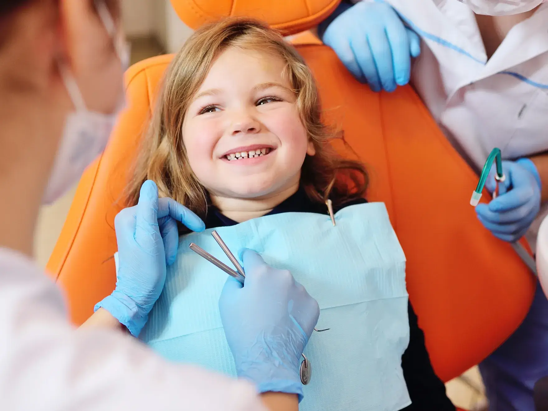 Smiling young girl sitting in an orange dental chair while a dentist wearing blue gloves prepares dental tools during a checkup.