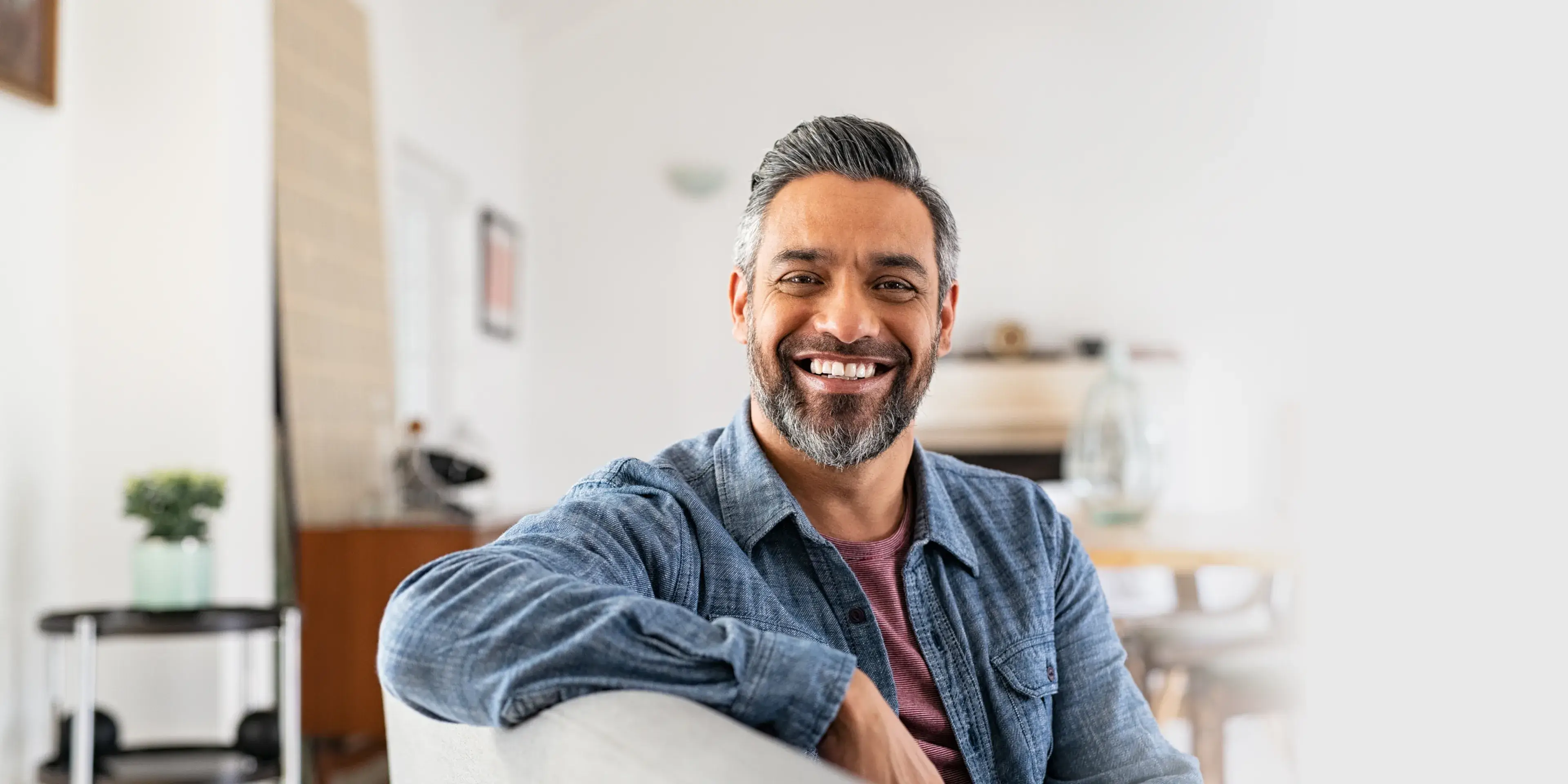 Smiling middle-aged man with salt-and-pepper hair and beard wearing a denim shirt sitting on a couch indoors.