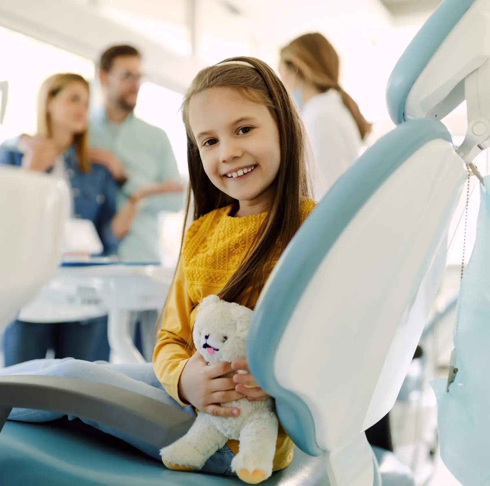 Smiling young girl in yellow shirt holding a stuffed animal while sitting in a dental chair with a dentist and parents blurred in the background.