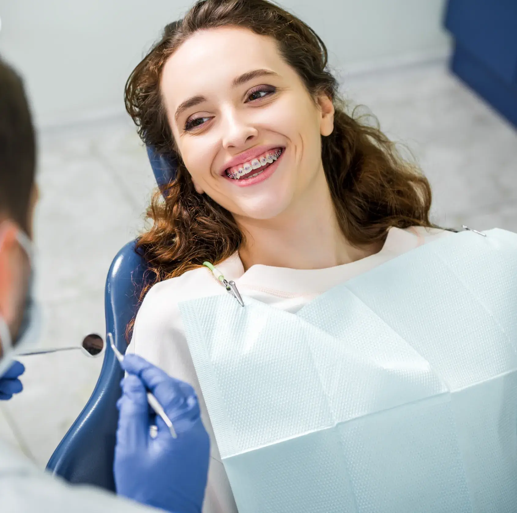 Woman with braces smiling in a dental chair during a checkup with a dentist wearing blue gloves.