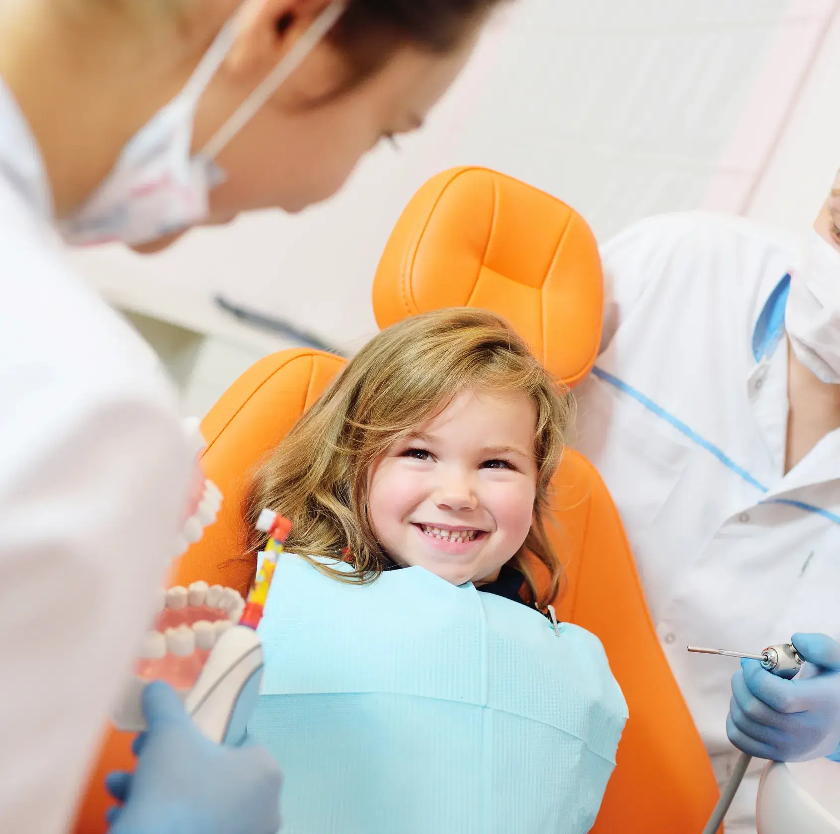 Smiling young girl in dental chair with dentist showing a toothbrush and a dental tool.