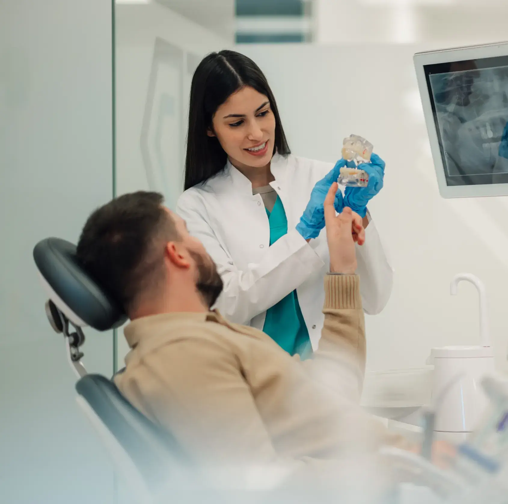 Dentist in a white coat and blue gloves explaining a dental model to a patient sitting in a dental chair.