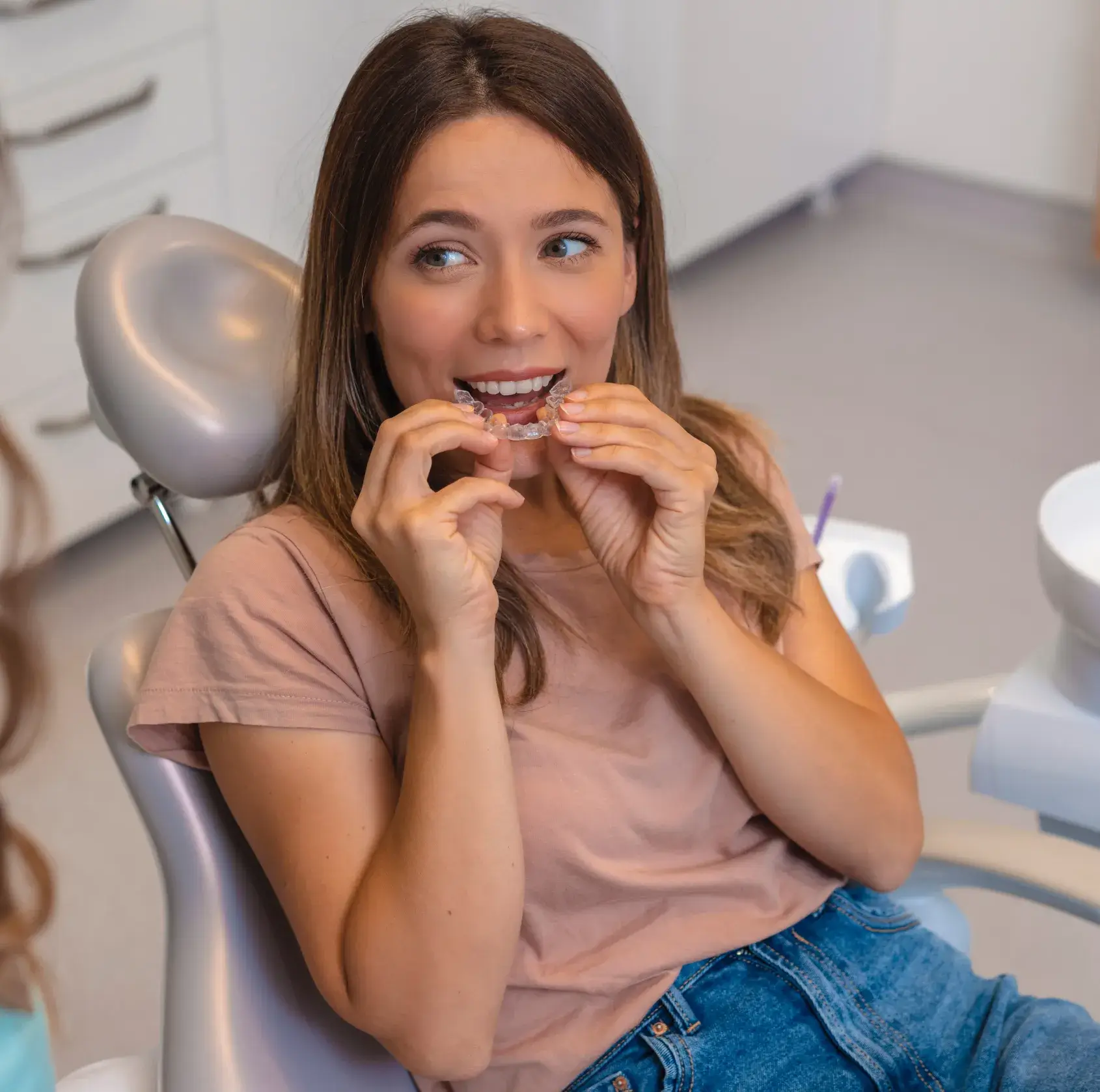 Young woman sitting in a dental chair about to insert clear Invisalign aligners on her teeth.