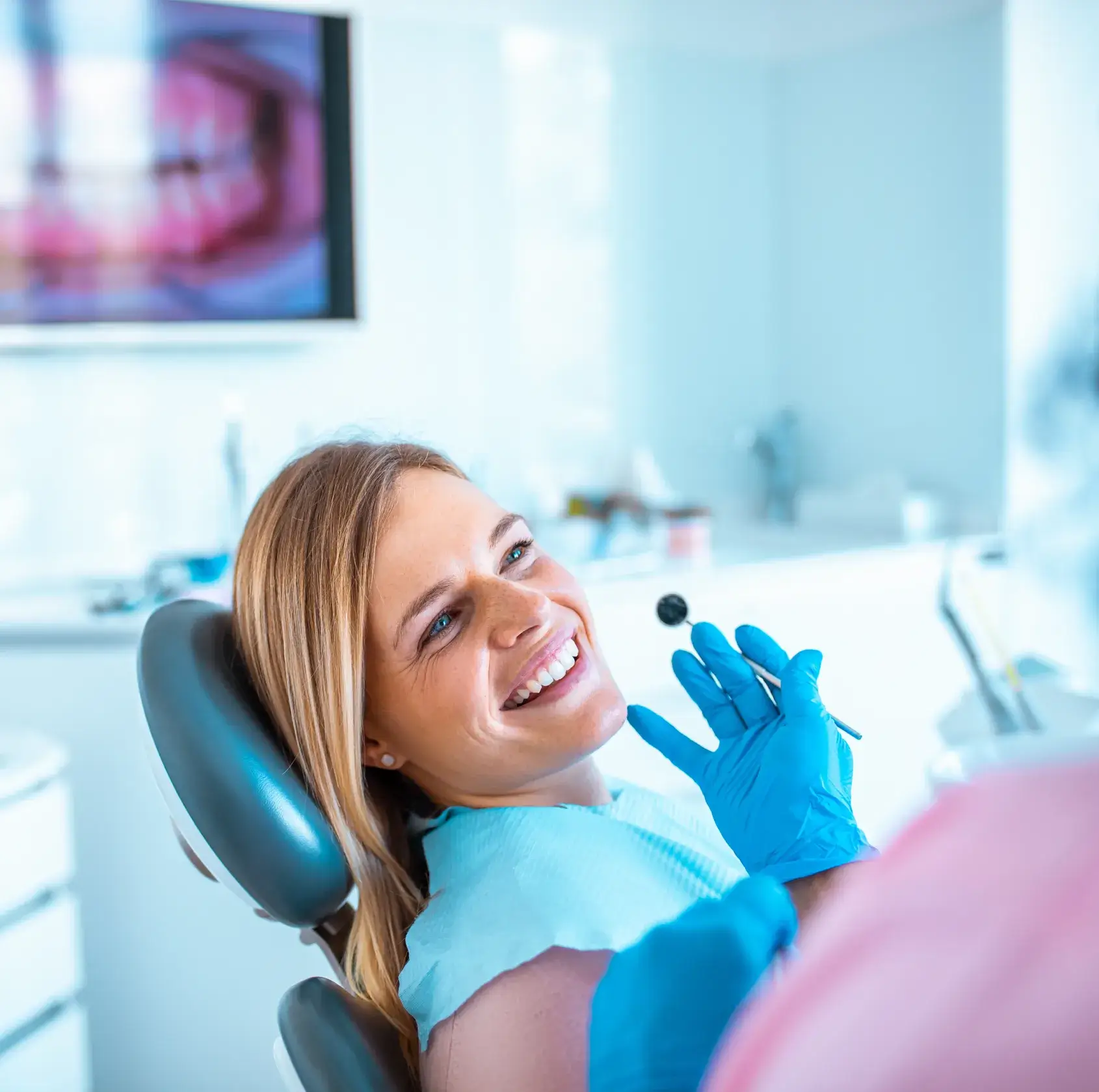 Smiling woman reclined in dental chair during a dental checkup with a gloved dentist holding a dental mirror.