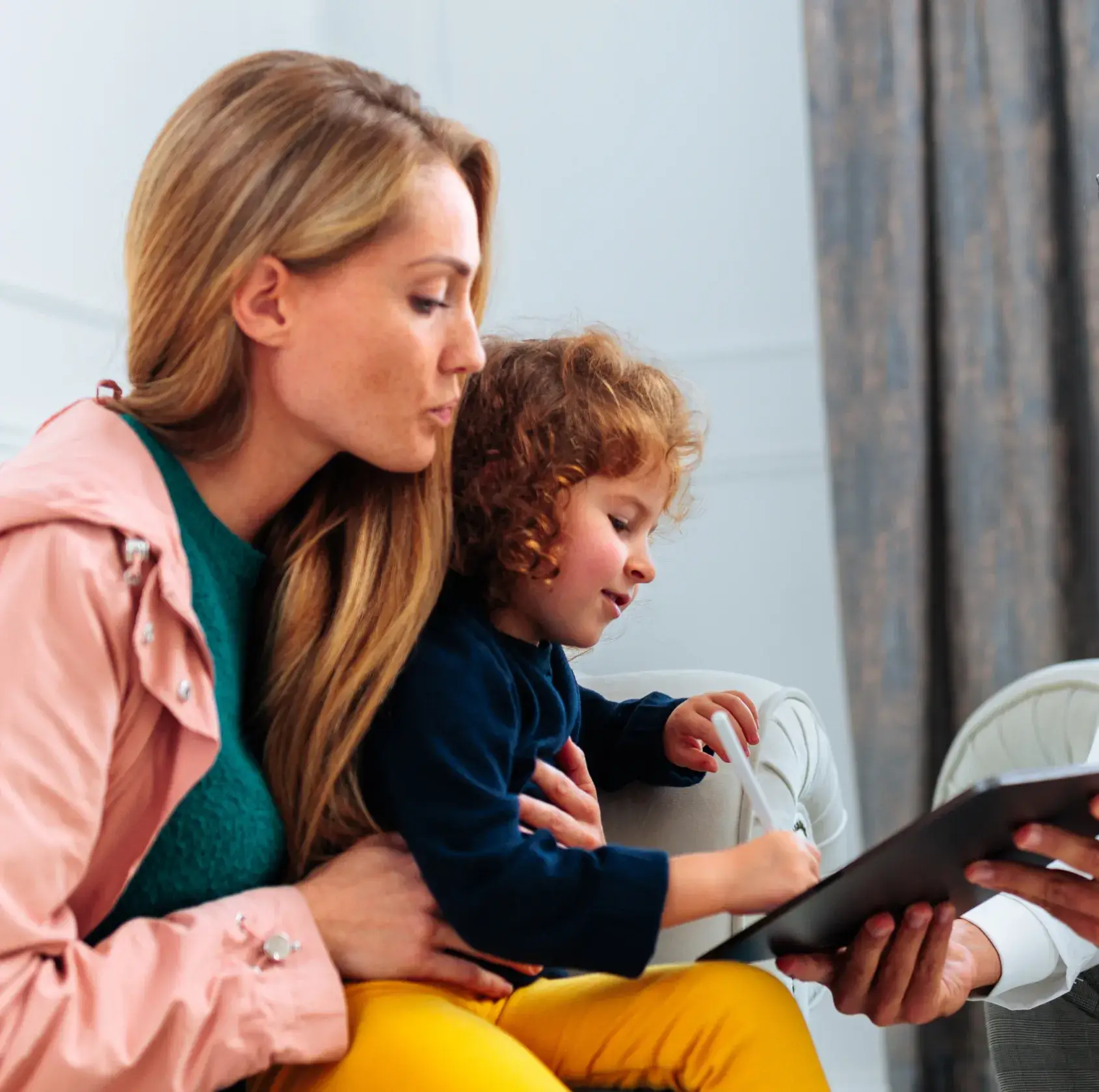 A woman gently holding a young child who is drawing on a tablet held by another person.