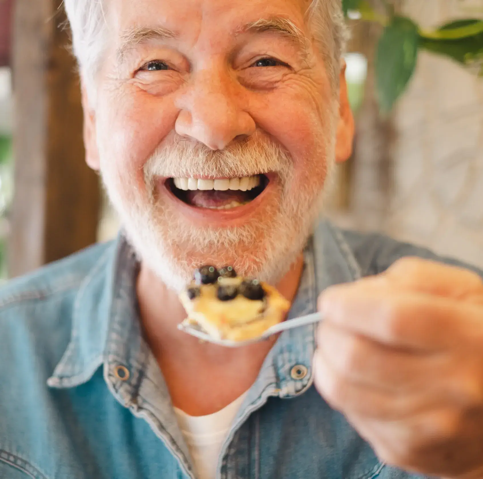 Smiling elderly man holding a spoon with a blueberry dessert close to his mouth.