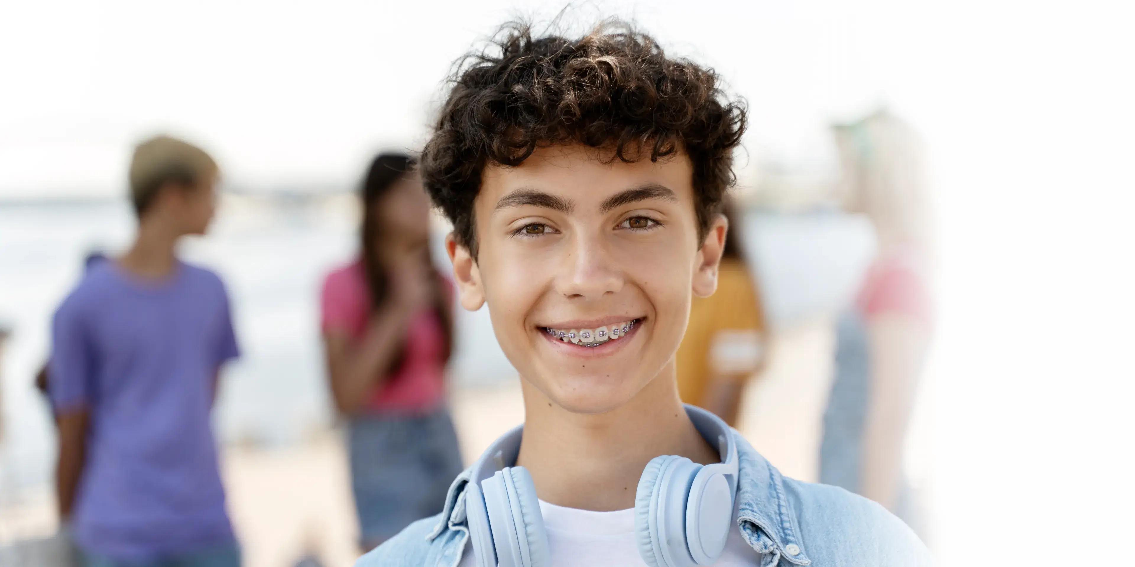 Teenage boy with curly hair and braces smiling, wearing headphones around his neck, with blurred friends in the background outdoors.