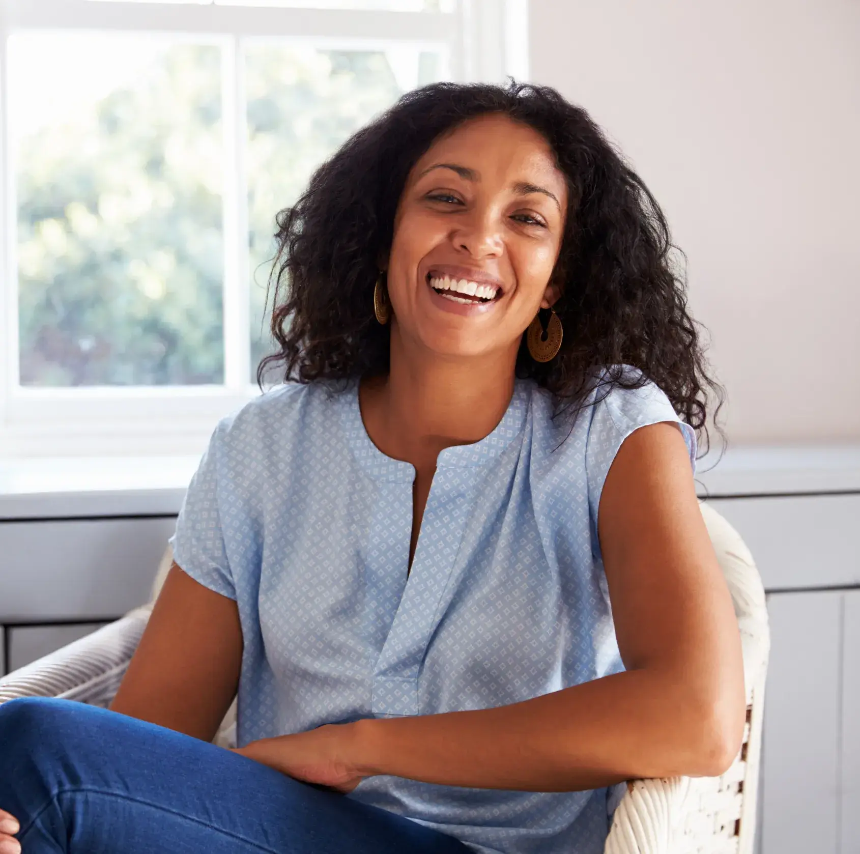 Smiling woman with curly hair wearing a light blue blouse and gold earrings sitting in a white wicker chair near a window.