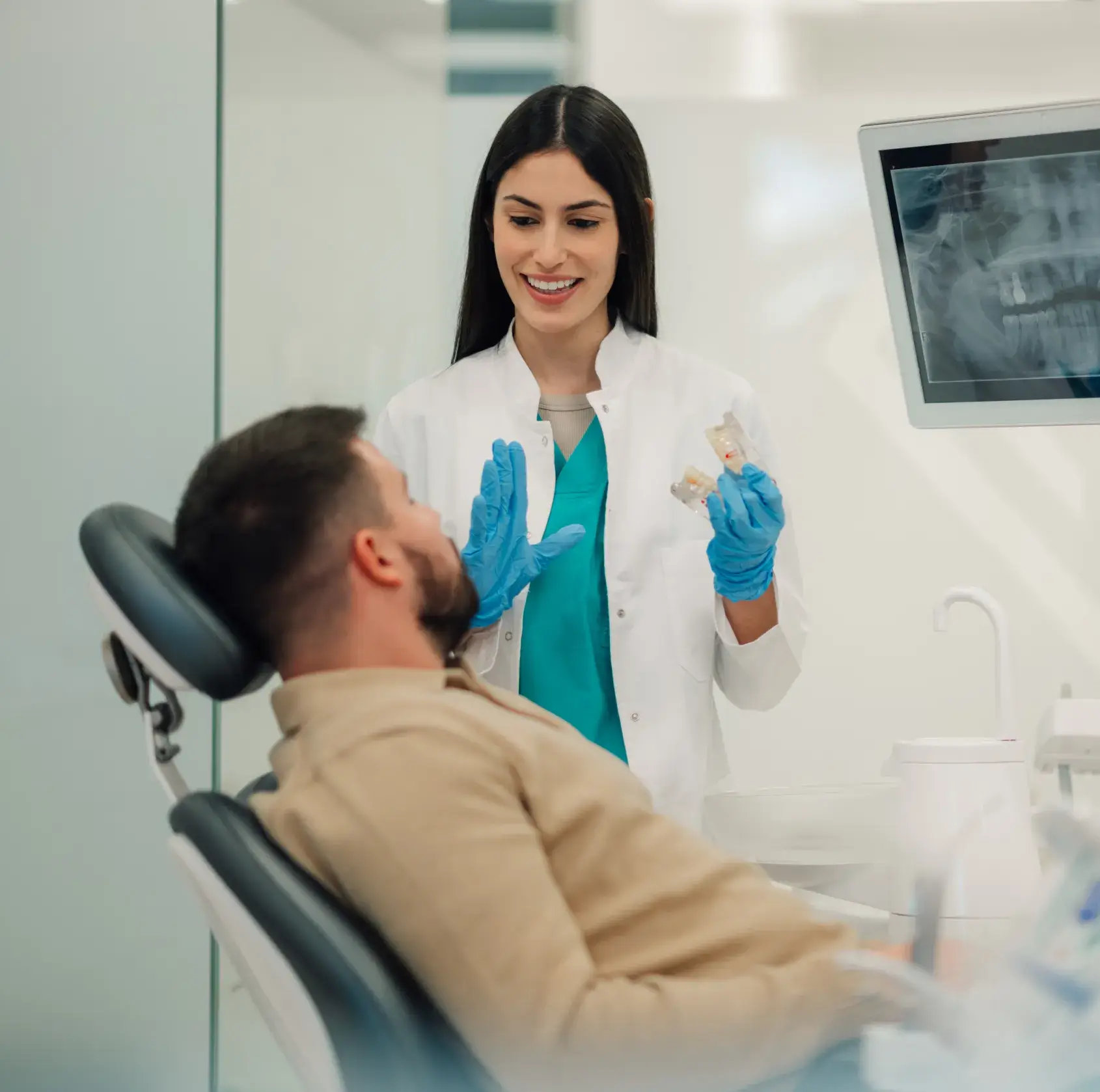 Dentist with blue gloves showing dental models to a seated male patient in a clinic.