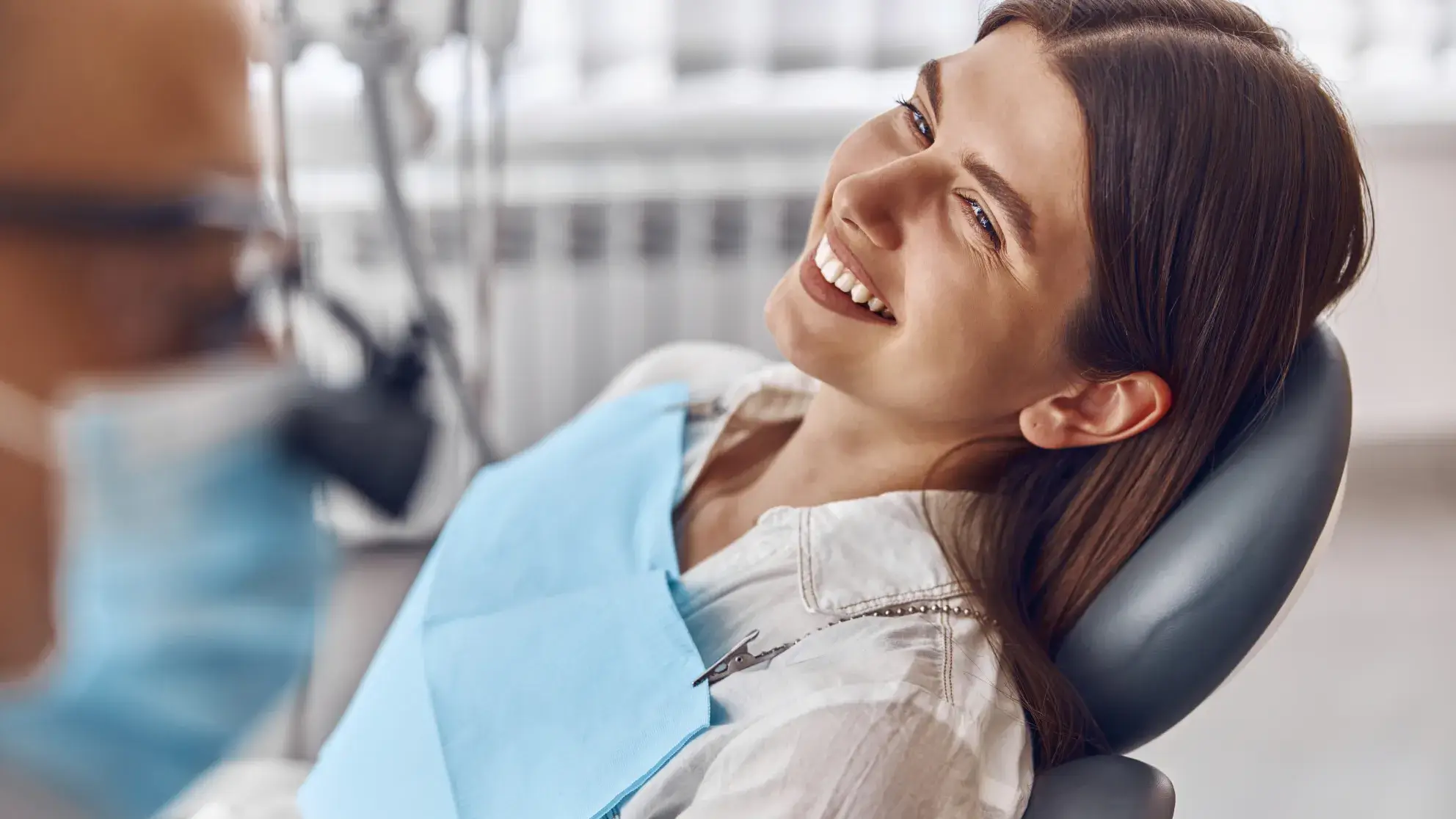 Smiling woman reclined in a dental chair wearing a blue bib, with a dentist partially visible wearing a face mask.