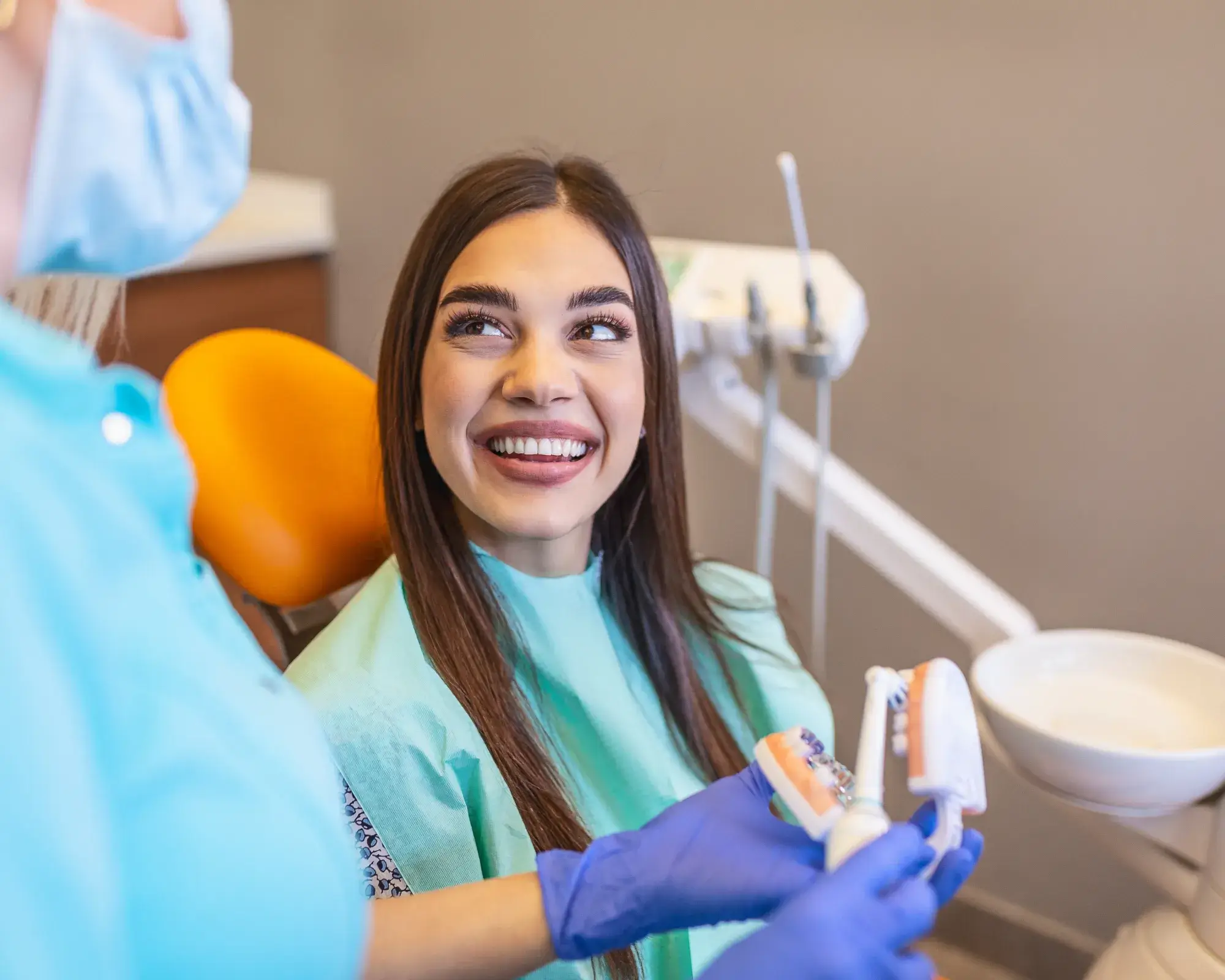 Smiling young woman in dental chair receiving oral hygiene instructions from masked dental professional holding a toothbrush and a dental model.