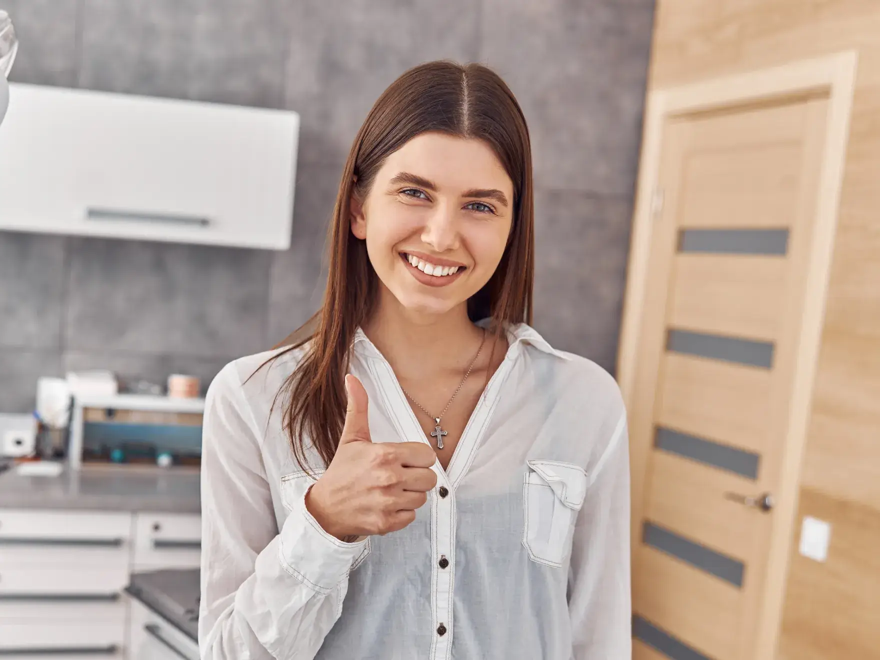 Smiling young woman in a white shirt showing thumbs up in a modern kitchen.