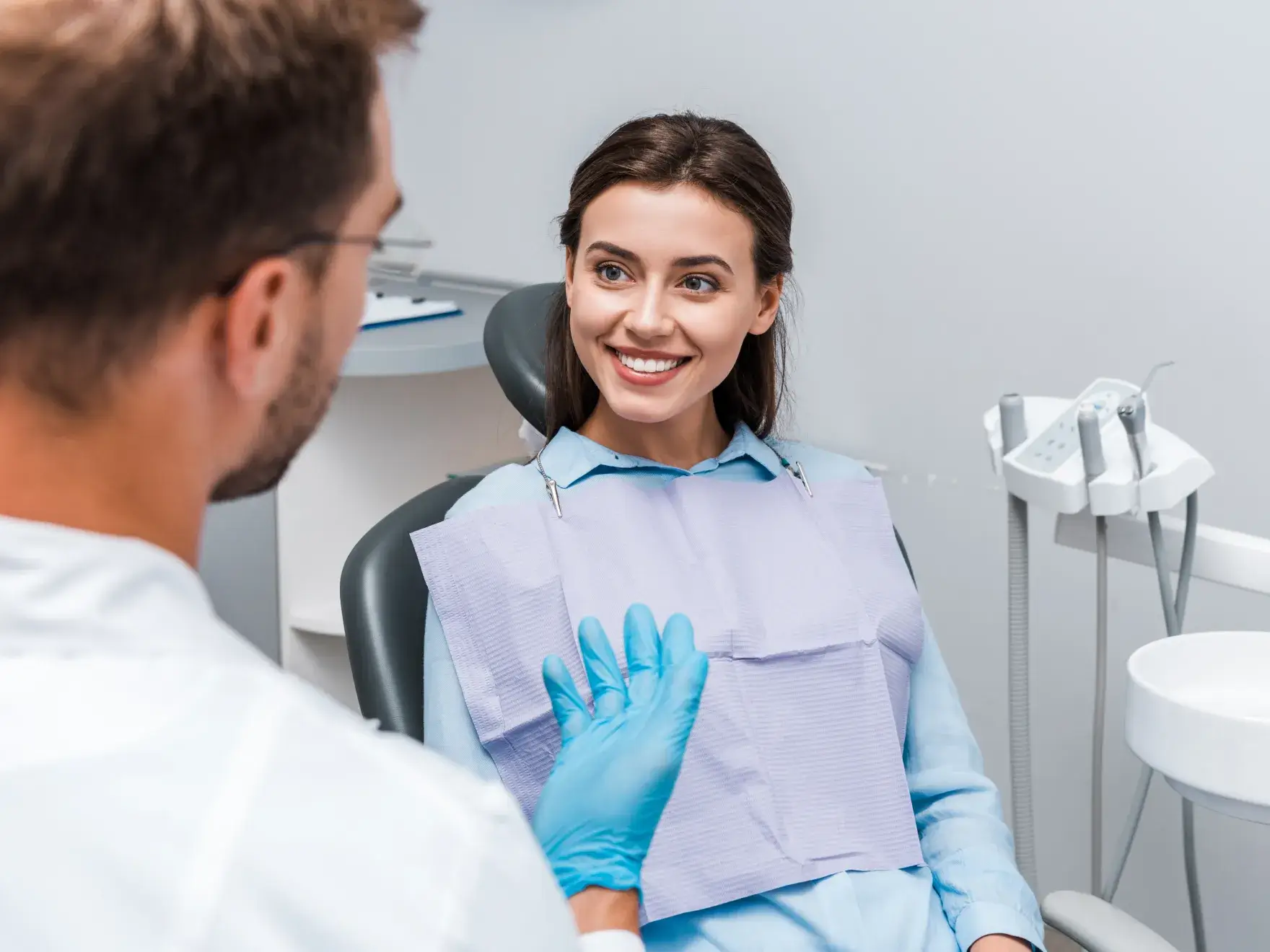 Smiling woman sitting in a dental chair, talking to a male dentist wearing blue gloves.