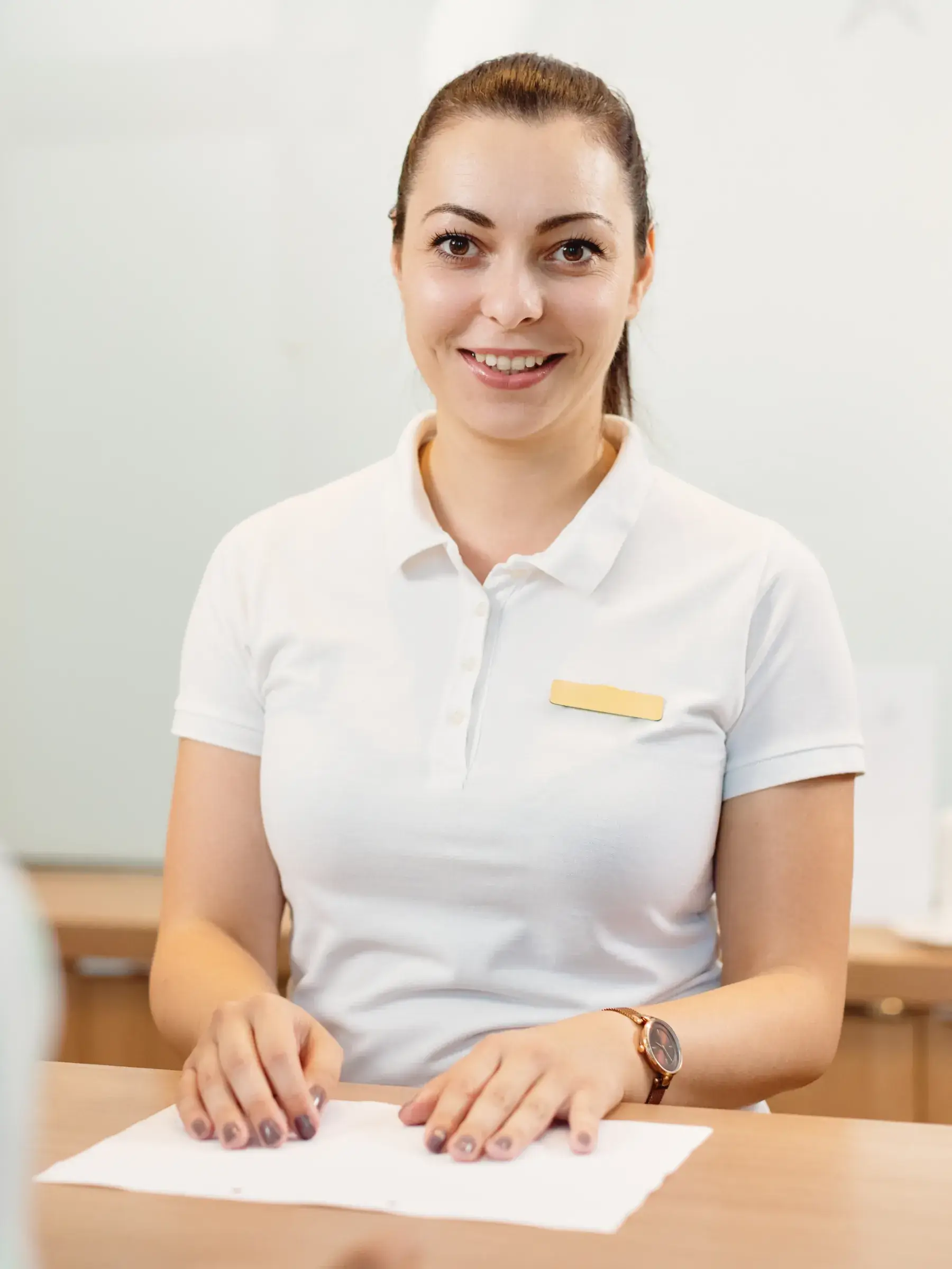 Smiling woman in white polo shirt sitting at a desk with papers, wearing a wristwatch and a blank name tag.