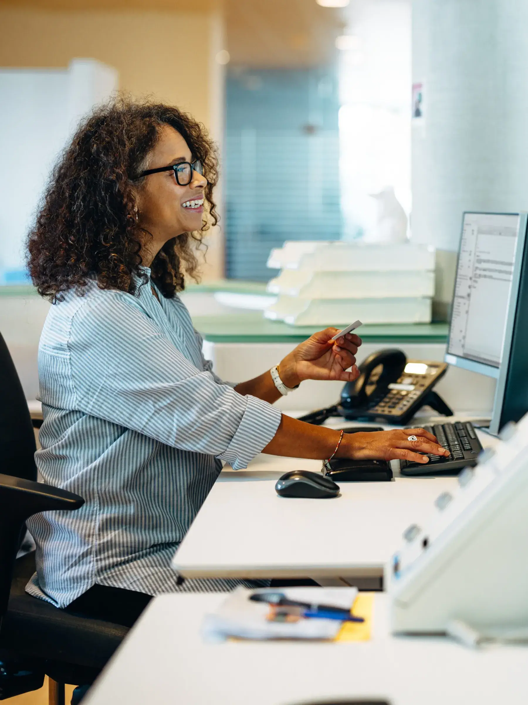 Smiling woman with curly hair and glasses working at a desk, using a computer keyboard and holding a card.