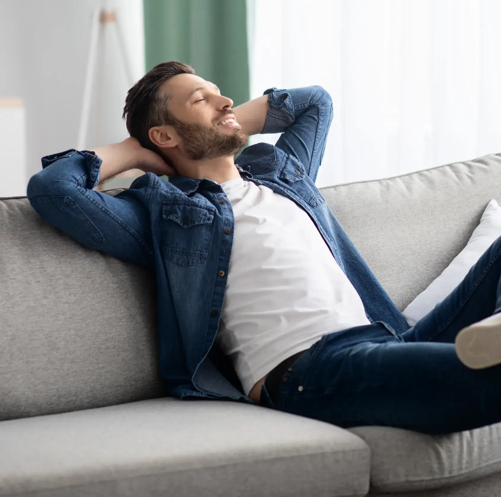 Smiling man in denim jacket reclining with hands behind head on gray sofa in a bright room.