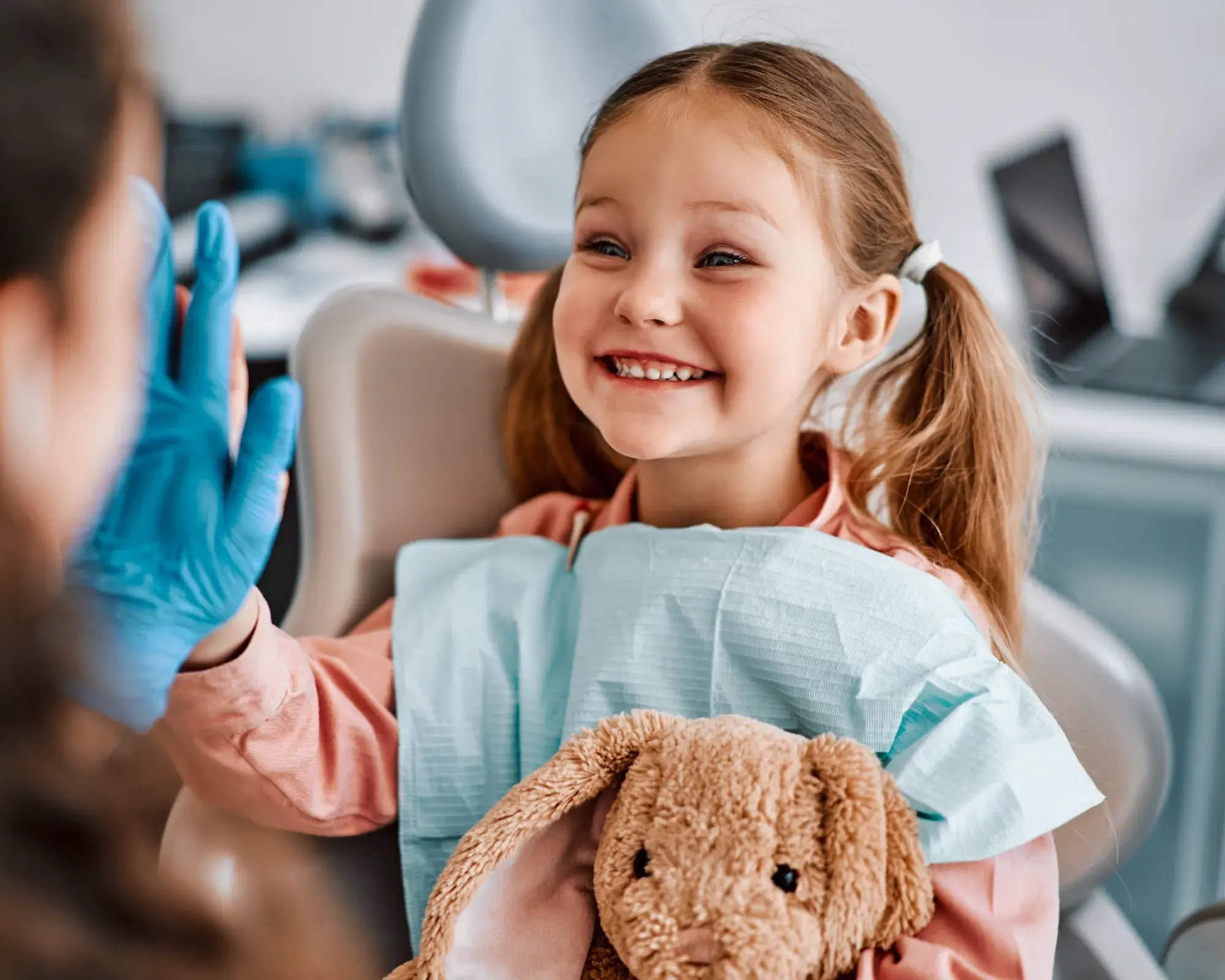 Smiling young girl in a dental chair giving a high five to a person wearing blue gloves, holding a stuffed bunny.