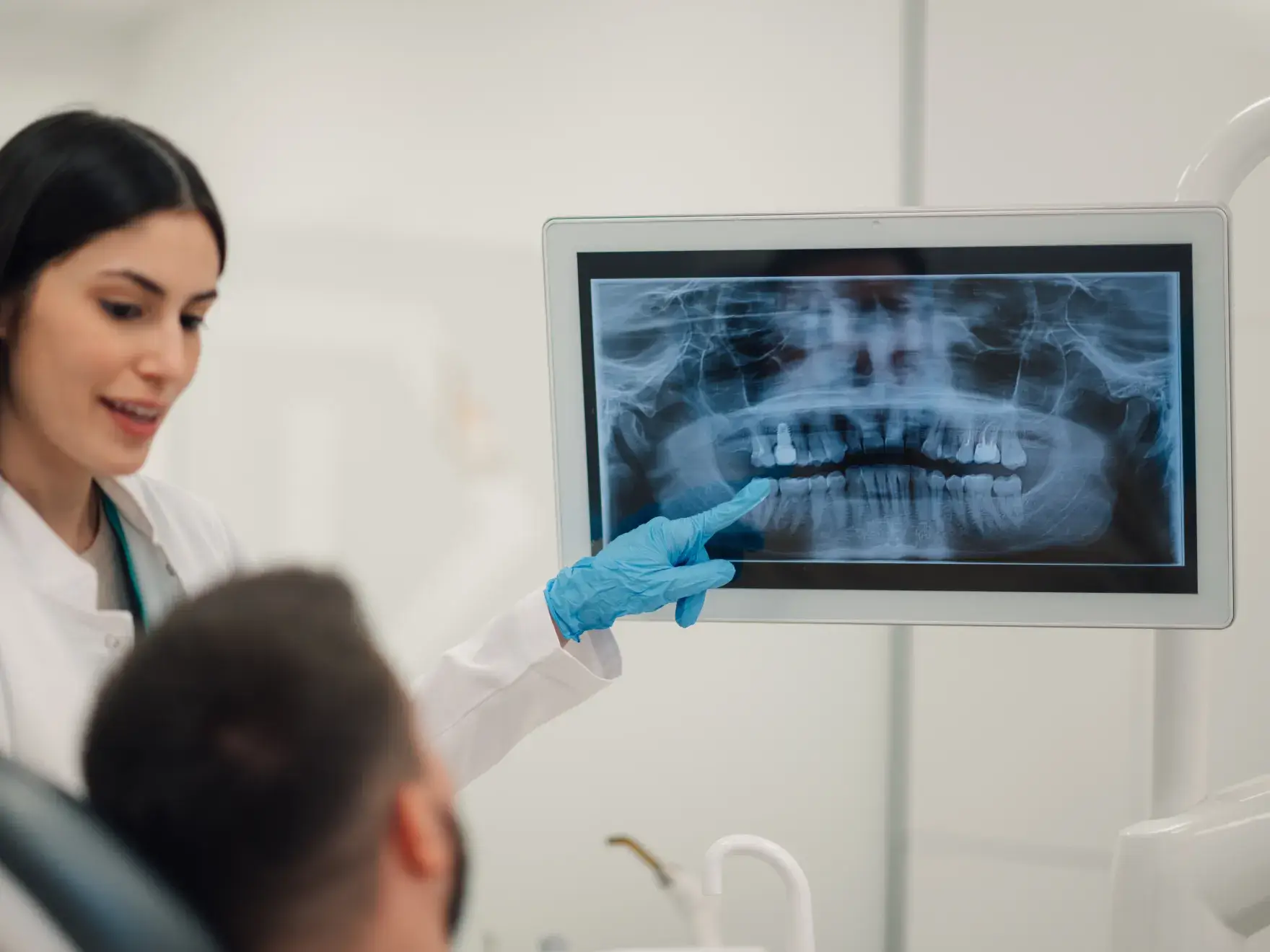 Dentist in white coat and blue gloves pointing at a dental X-ray on a monitor while explaining to a seated patient.