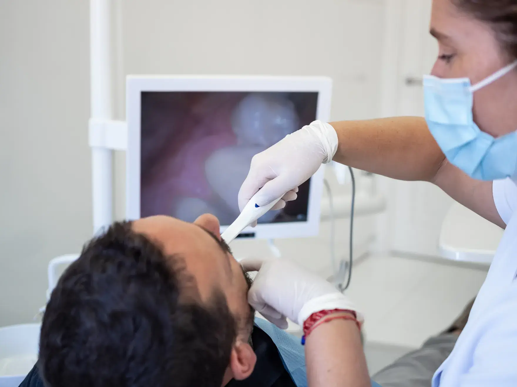 Dentist wearing gloves and a mask using an intraoral camera to examine a male patient's mouth with an image displayed on a nearby monitor.