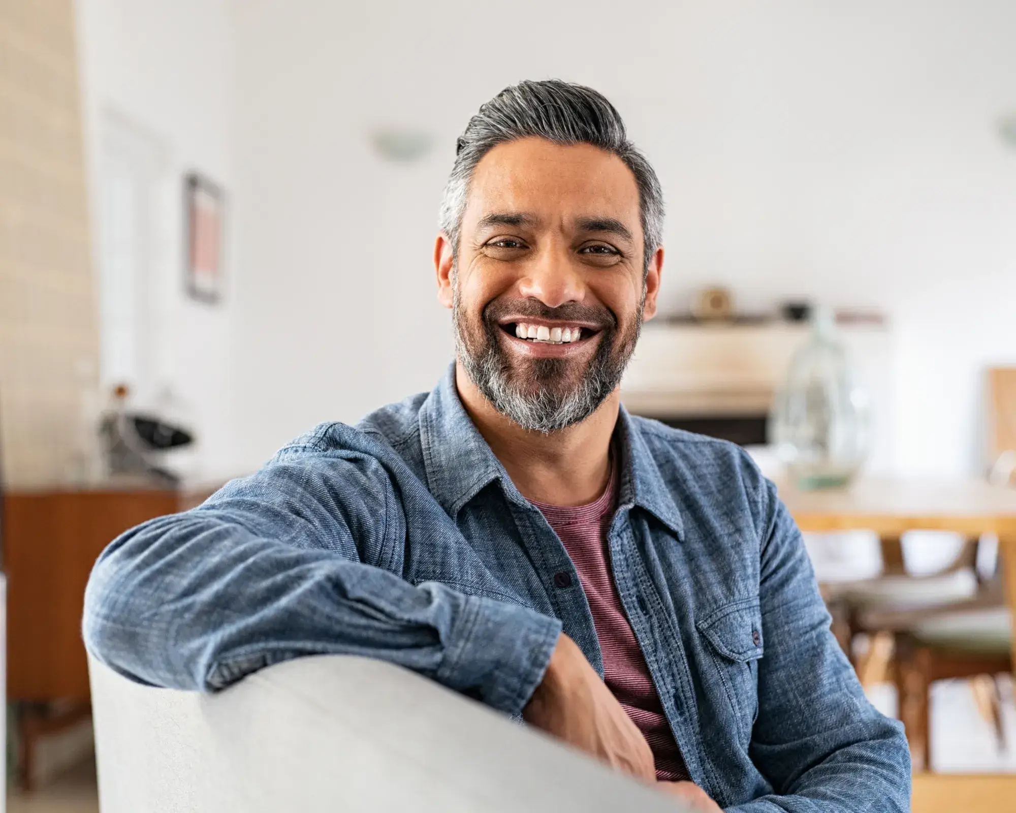 Smiling middle-aged man with gray hair and beard sitting on a couch in a bright living room.