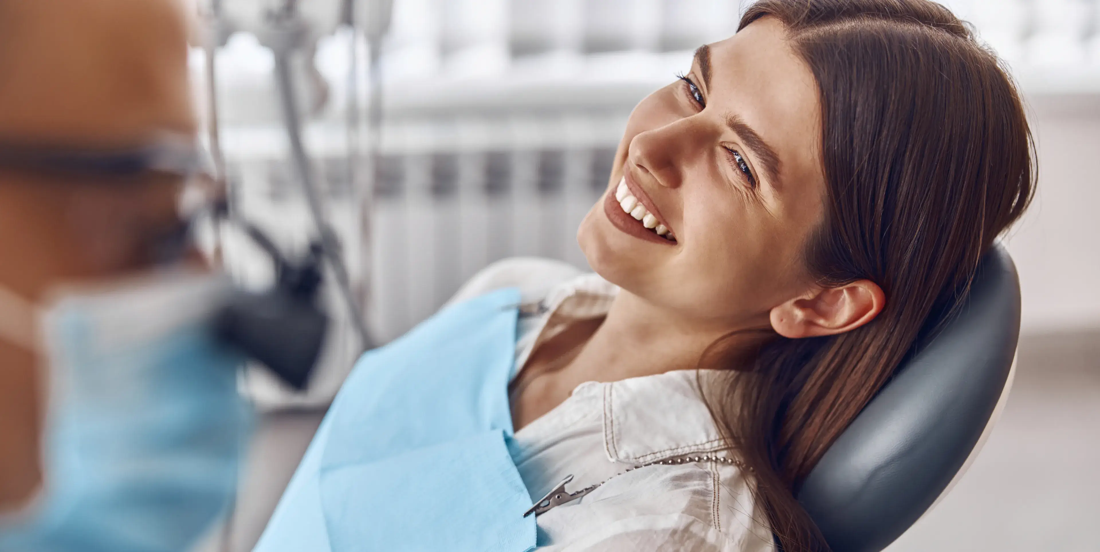 Smiling woman reclining in a dental chair wearing a blue bib with a dentist in a mask blurred in the foreground.