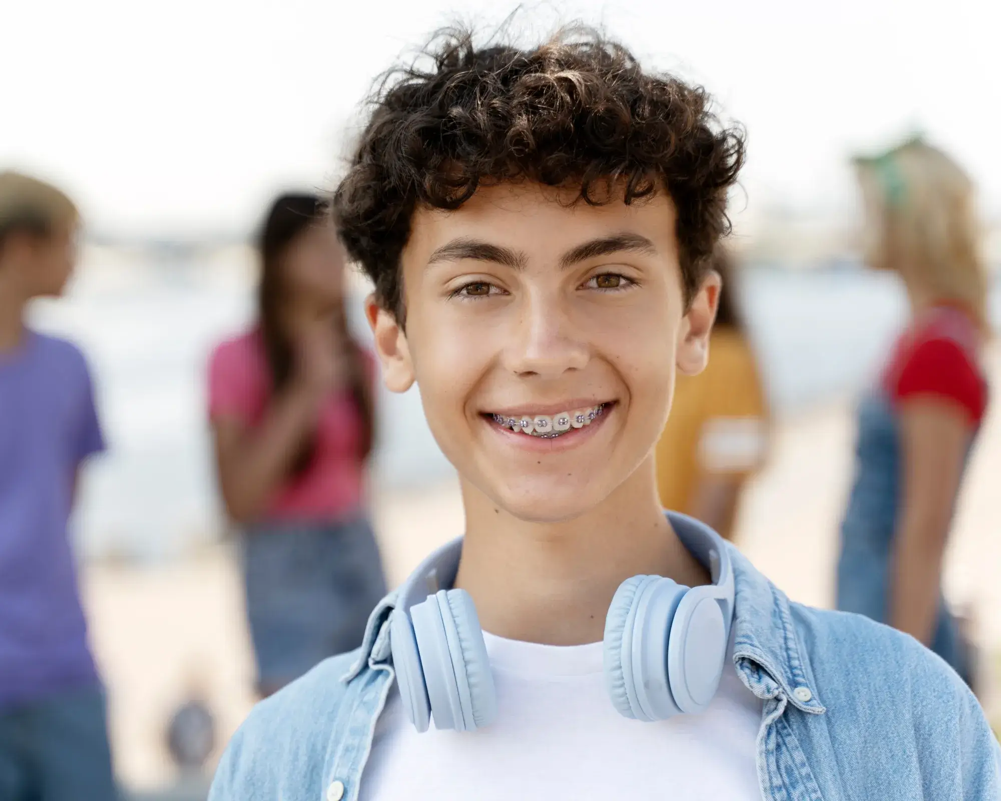 Teenage boy with curly hair, braces, and light blue headphones around his neck smiling at the camera with three blurred people in the background.