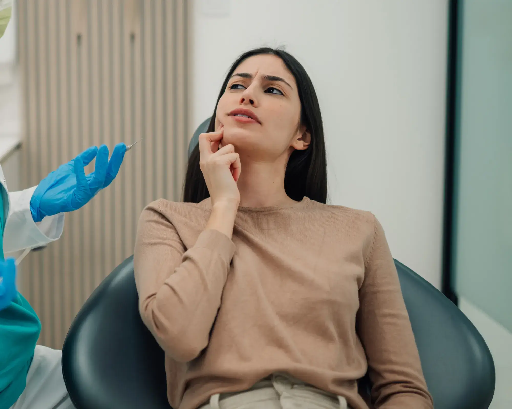 Young woman sitting in a dental chair looking concerned while a dentist wearing blue gloves holds a syringe nearby.