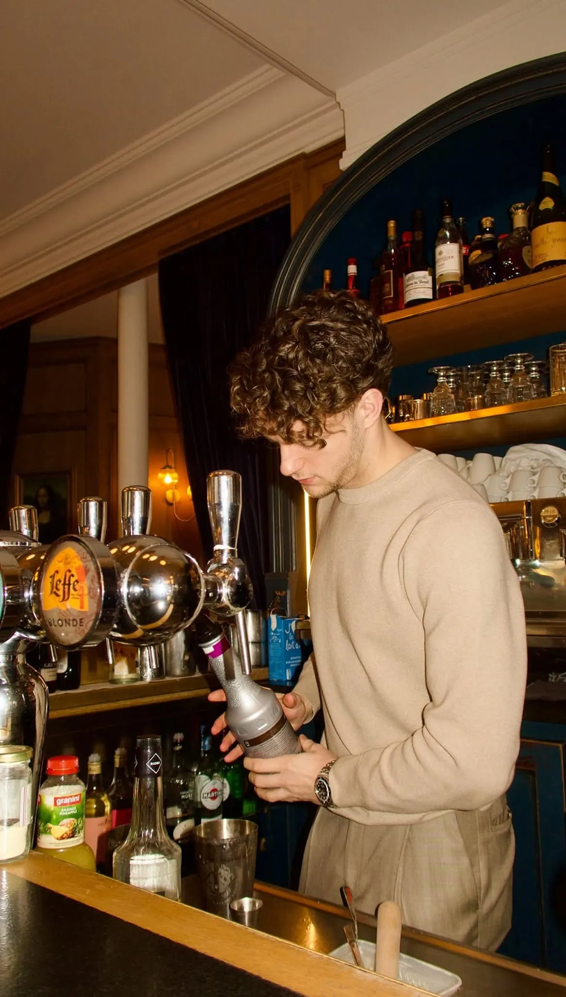 Young man with curly hair in beige sweater pouring a drink behind a bar with bottles and taps.
