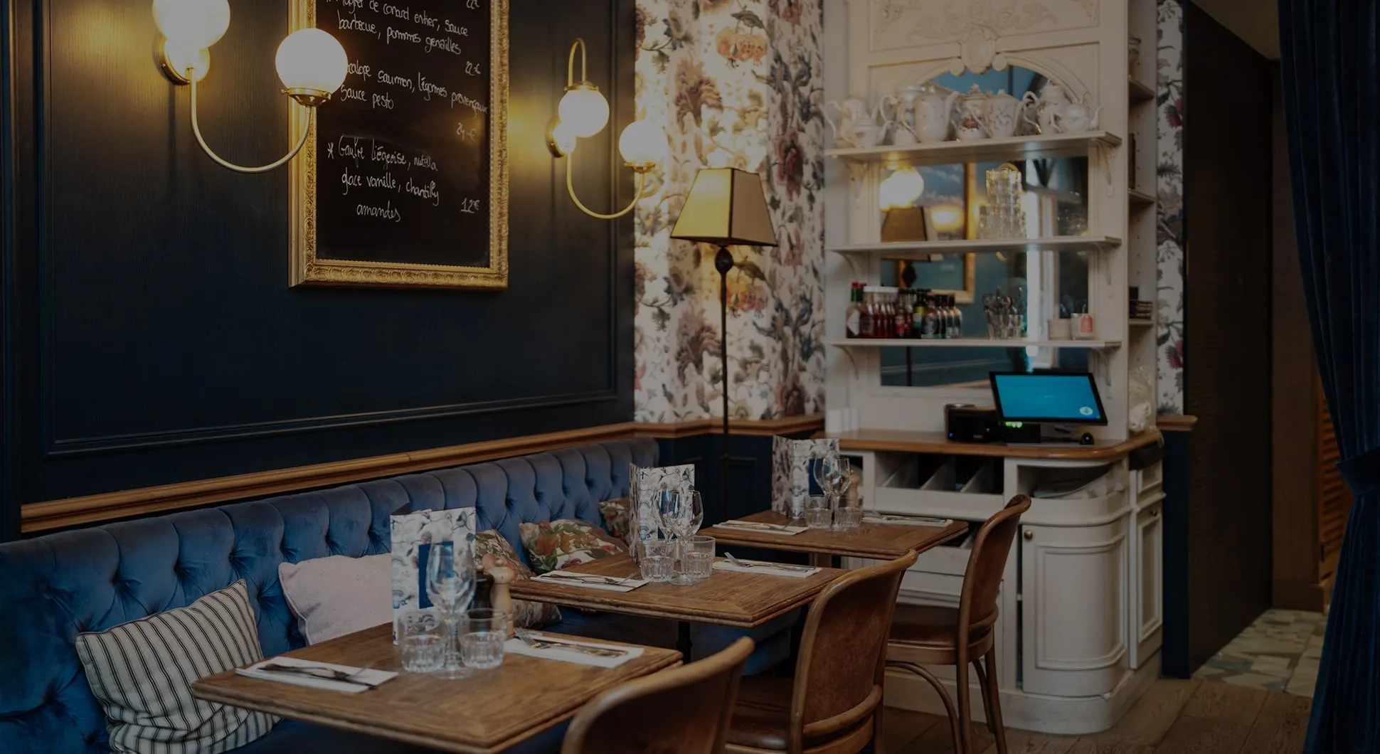 Cozy restaurant interior with wooden tables, cushioned blue bench seating, floral wallpaper, and warm lighting.