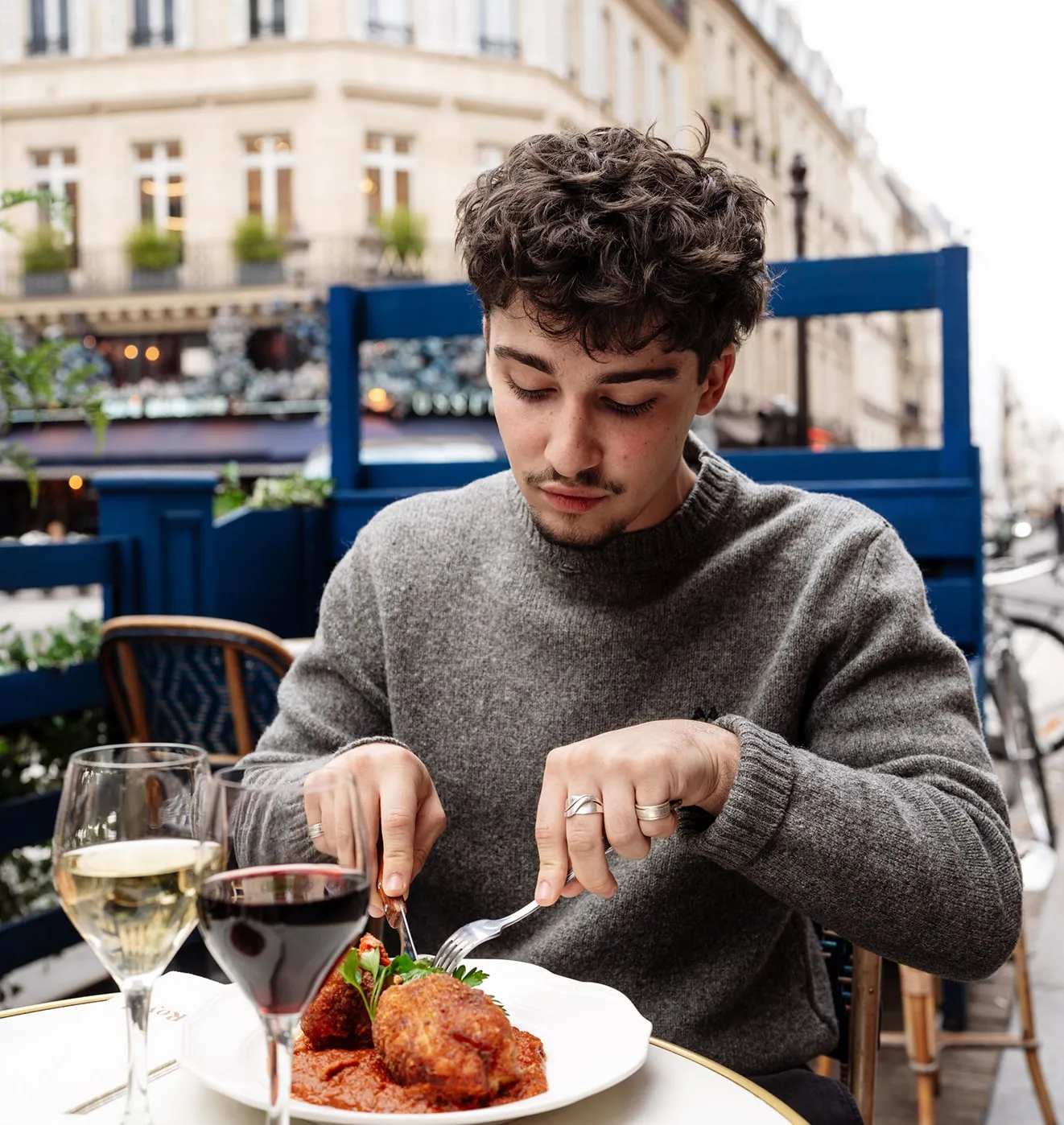 Young man in a grey sweater dining outdoors, cutting food on a white plate with red sauce, with glasses of red and white wine on the table.