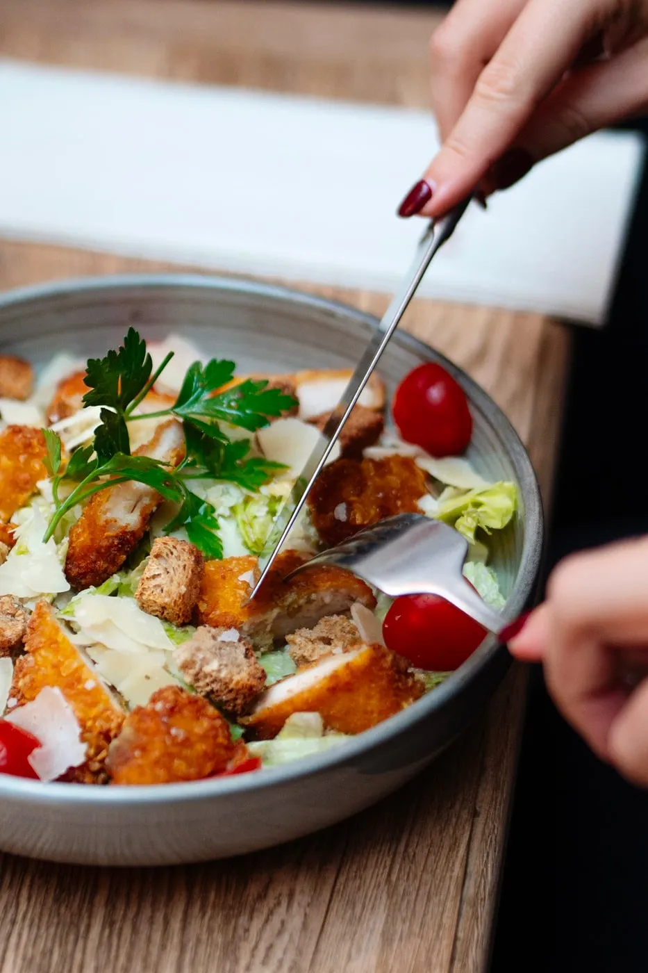 Close-up of a person cutting crispy chicken pieces in a Caesar salad bowl with croutons, cherry tomatoes, and parsley garnish.
