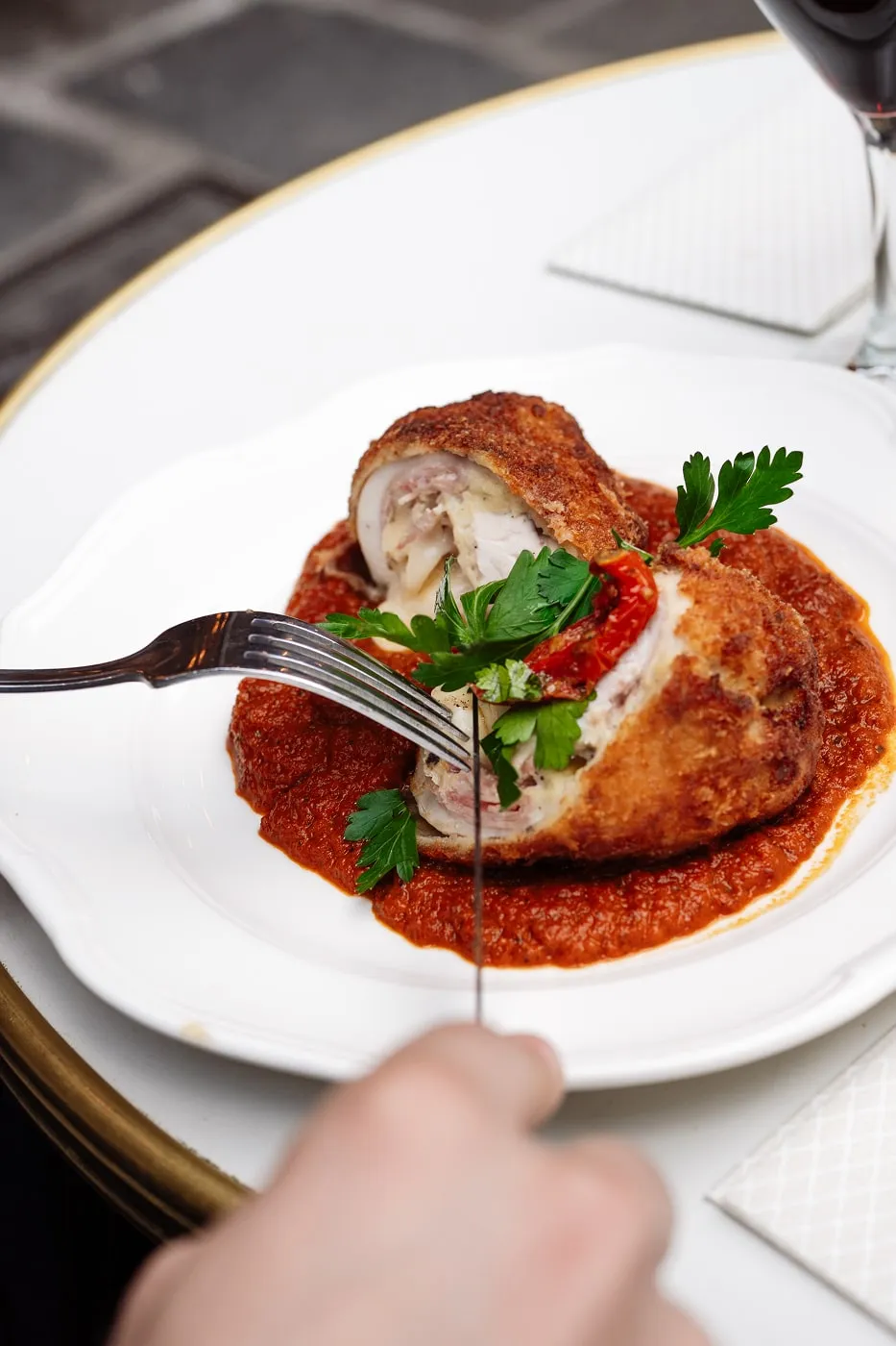 Hand cutting into breaded chicken stuffed with cheese and herbs, served on marinara sauce with parsley garnish on white plate.