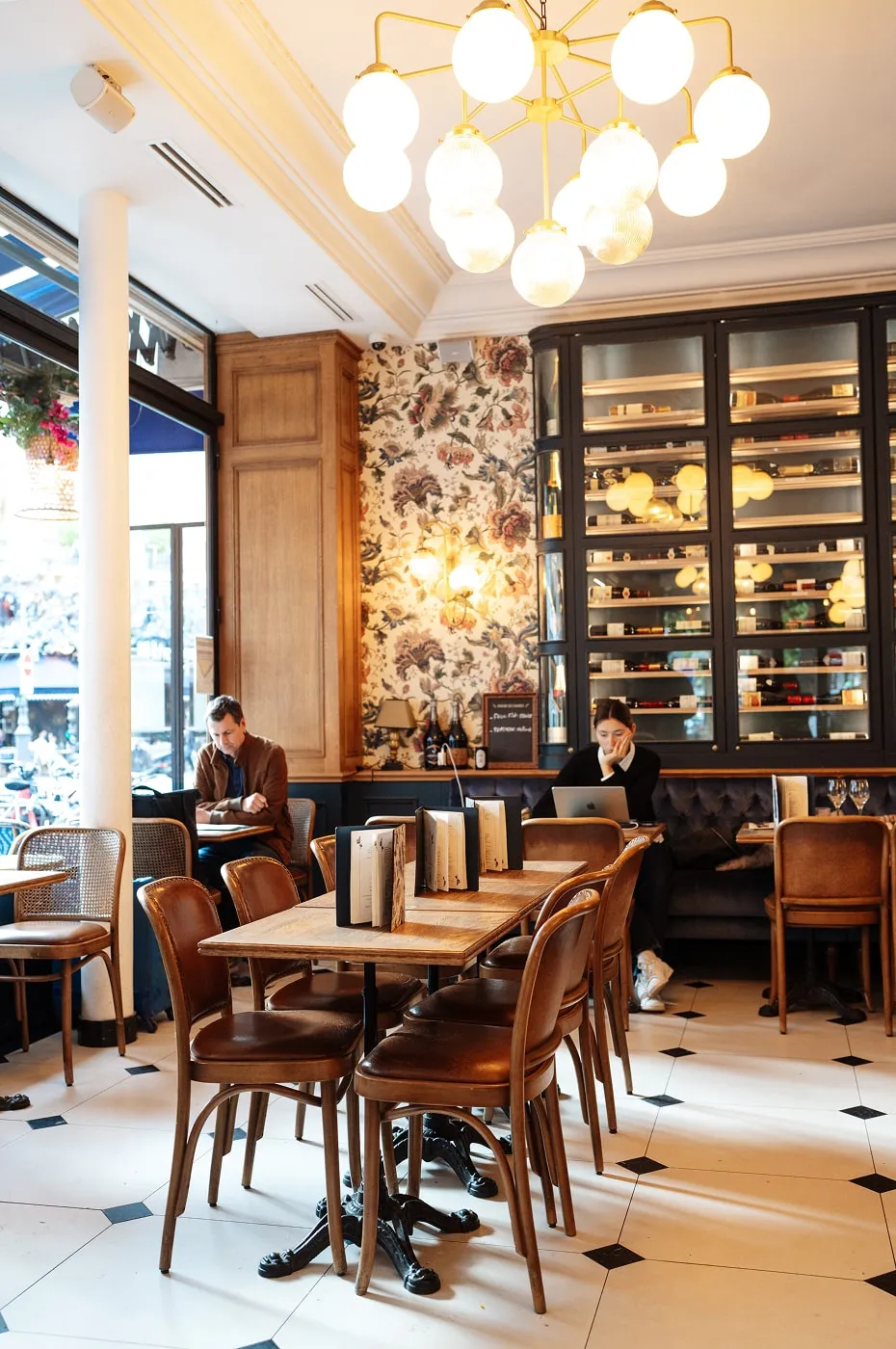 Cozy cafe interior with wooden tables and chairs, floral wallpaper, wine cabinet, and two people working—one on a laptop and one reading.